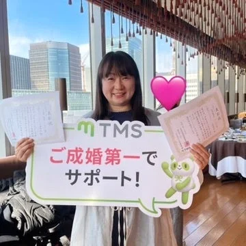 笑顔の女性が看板と証明書、「ご成婚第一でサポート！」