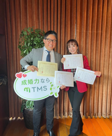 結婚相談所mTMSのロゴ入り看板と複数の表彰状を手に笑顔で立つ男女