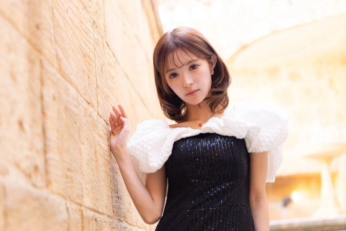 黒いスパンコールのドレスと白いパフスリーブを着用した若い女性が、明るい色の石壁にもたれかかってポーズをとっています。