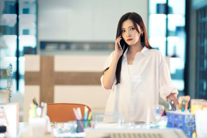 白いシャツを着た若い女性がオフィスのような場所で電話をしている様子です。彼女は真剣な表情で、長く黒い髪が特徴的です。