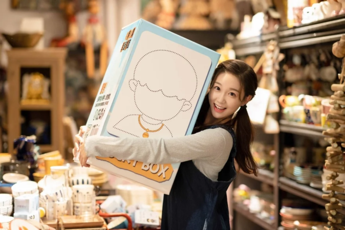 笑顔の若い女性が、頭部のイラストが描かれた大きな箱を抱えて店内に立っている。楽しげな表情でカメラを見つめている。