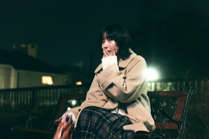 夜の屋外でベンチに座る若い女性のポートレート。ベージュのコートとチェック柄のスカートを着用し、少し驚いたような、あるいは物思いにふけるような表情で、片手を口元に当てています。背景は暗く、街灯の光が印象的です。