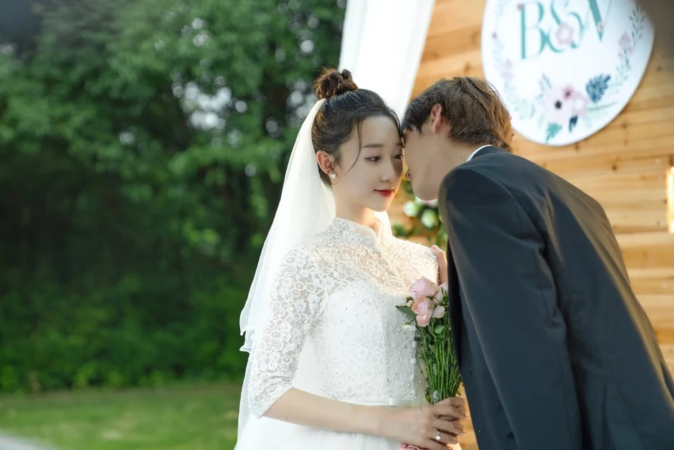 屋外の結婚式で、新郎が新婦に優しく寄り添うロマンチックな瞬間を捉えた画像です。新婦は白いウェディングドレスを着てブーケを持っています。