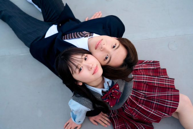 制服姿の若い男女が寄り添い、地面に寝転がってカメラを見上げているポートレート写真です。二人は優しい笑顔を浮かべ、青春の甘く温かい瞬間が捉えられています。