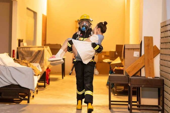 消防士が完全装備で女性を抱きかかえ、救助している様子。散らかった室内が背景にあり、緊急時の救助活動を描写しています。消防士の勇敢さと女性の安全が際立つ一枚です。