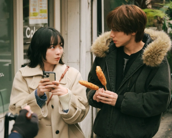 冬の装いをした若い男女が街角でくつろいでいる様子。男性は串に刺さった揚げ物を持ち、女性はスマートフォンを手に彼を見つめている。日常的な一コマを捉えた写真。