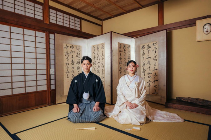 日本の伝統的な家屋で、紋付袴と白無垢を着用した新郎新婦が畳に正座している。背景には書道の施された屏風があり、厳かで美しい和装の結婚写真を捉えています。