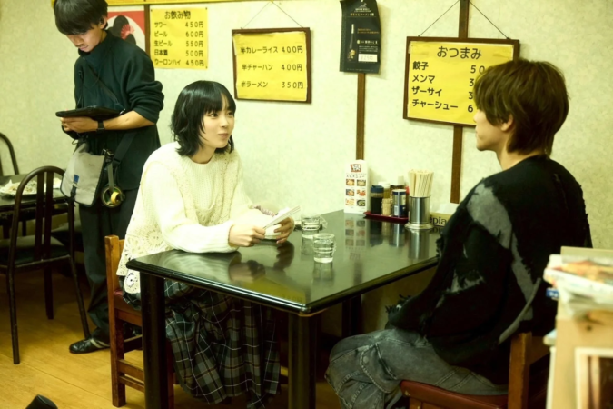 日本の食堂のような場所で、男女二人がテーブルを挟んで向かい合って座り、会話している様子。壁には飲み物やおつまみ、食事のメニューが貼られており、店員らしき男性が近くに立っている。