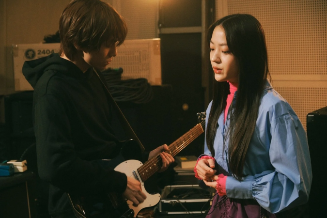 スタジオで男性がエレキギターを演奏し、隣で女性がその様子を見守っている場面です。二人の若者が音楽に集中しているような雰囲気が伝わります。