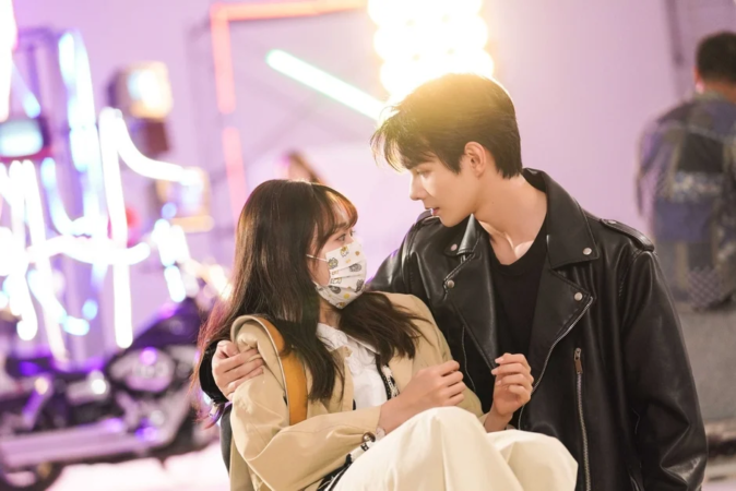 ネオンライトが輝く屋内で、若いカップルが互いに見つめ合っているロマンチックなシーン。男性はレザージャケットを着用し、女性は猫柄のマスクを着けている。