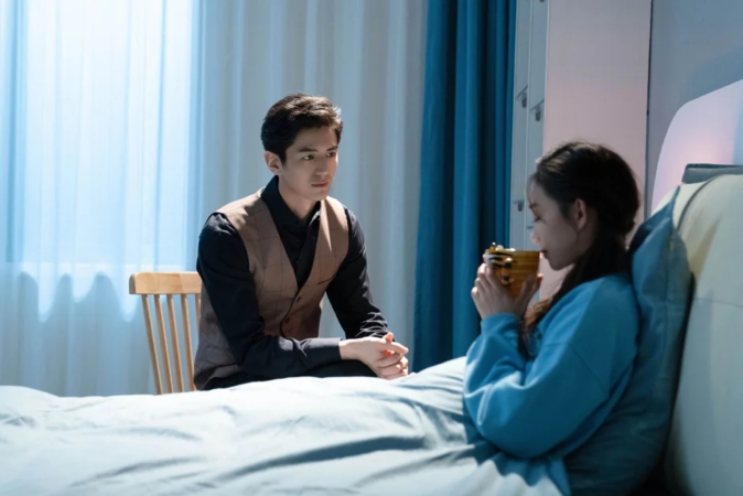 ベッドで横になっている女性がカップで飲み物を飲んでおり、その傍らで男性が椅子に座って女性を見守っている様子。室内で二人が向き合っている、穏やかな一場面です。