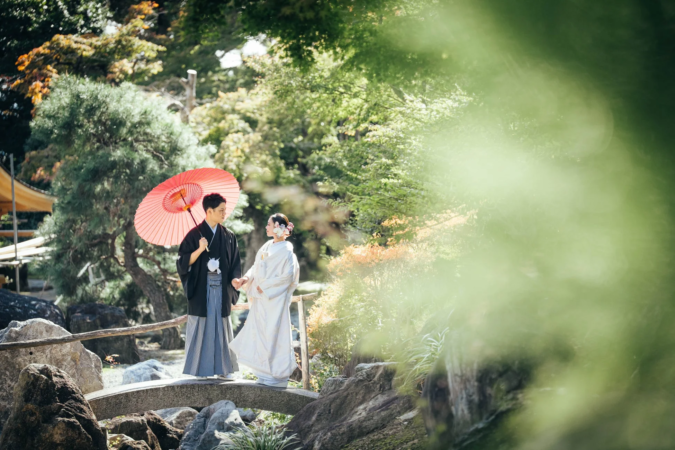 美しい日本庭園の石橋を、和装姿のカップルが手を取り合って歩いています。新郎は赤い和傘を差し、新婦は白無垢をまとっています。柔らかな日差しが差し込む、穏やかでロマンチックな婚礼の情景です。