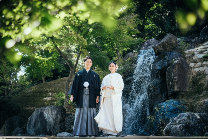 日本の伝統的な結婚衣装をまとった新郎新婦が、緑豊かな庭園の滝の前で笑顔で立つ姿を捉えた一枚。和やかな雰囲気で、結婚式の前撮り写真と思われる。