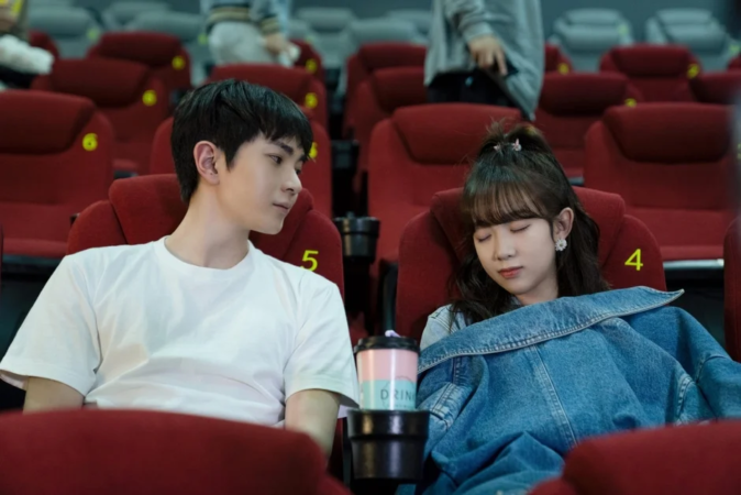 映画館で、白いTシャツを着た男性が隣でデニムジャケットを羽織って眠っている女性を見つめている様子です。二人の間には飲み物が置かれ、リラックスした雰囲気が漂っています。