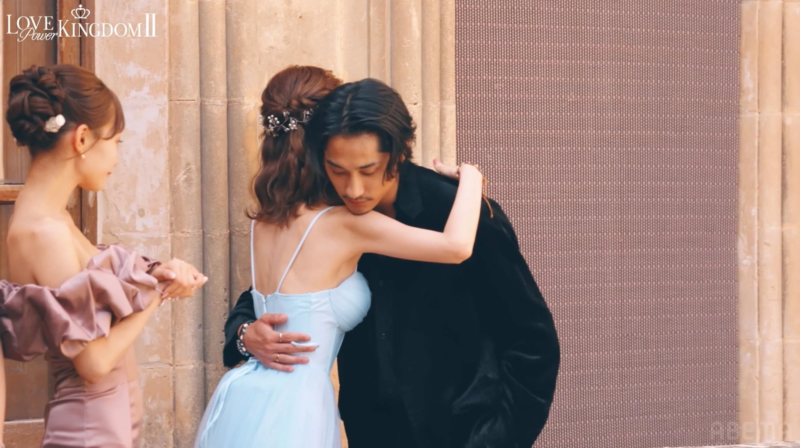 男性が水色のドレスの女性を抱きしめており、別の女性がその様子を見つめているロマンチックなシーン