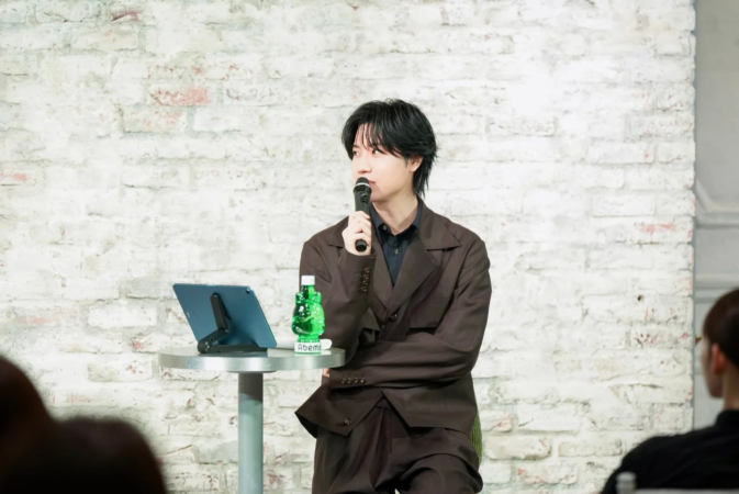 白いレンガ壁を背景に、ダークブラウンのスーツを着た男性がマイクを持って話しています