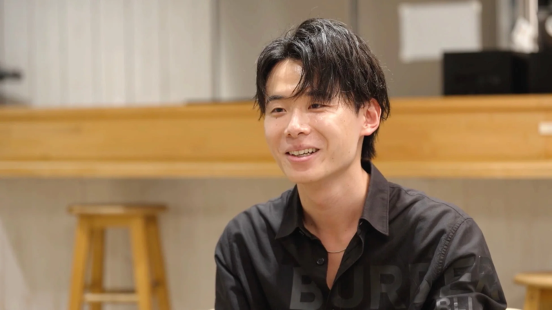 黒いシャツを着た若いアジア人男性が、明るい笑顔で左側を見ています。屋内での自然な表情のポートレートで、親しみやすい雰囲気が感じられます。