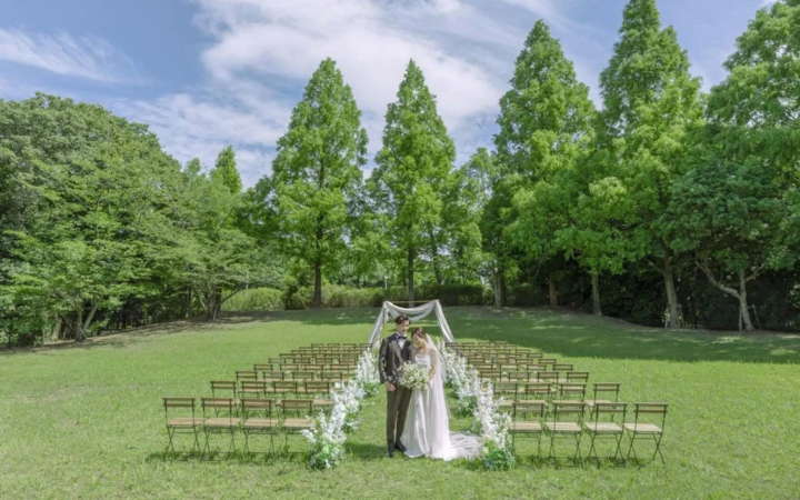 緑豊かな芝生広場で、白い花と椅子が並べられたバージンロードの先に新郎新婦が立つ、開放的なアウトドアウェディングの風景です。