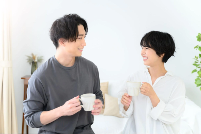 明るい部屋で、マグカップを持った男女が笑顔で向かい合い、リラックスした雰囲気で会話を楽しんでいる様子です。
