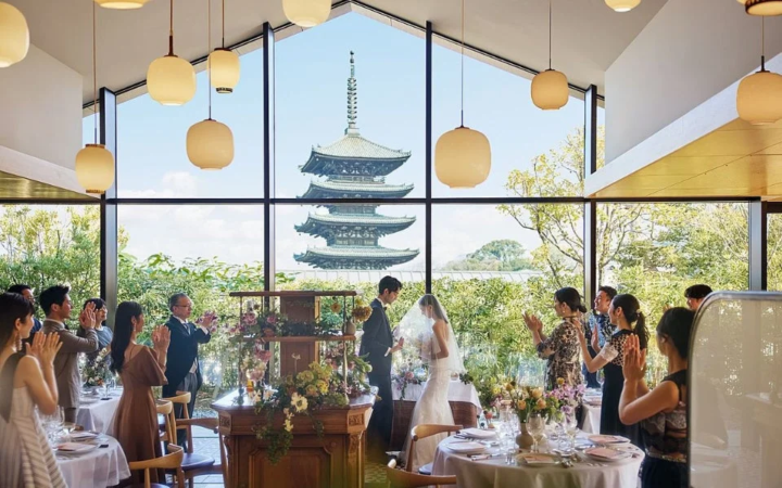 大きな窓から五重塔を望むモダンな会場で、新郎新婦がゲストに囲まれて結婚式を挙げている様子。