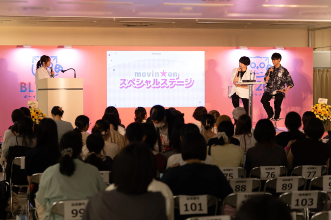 「movin'on スペシャルステージ」と表示されたスクリーンがあるステージで、女性と2人の男性が登壇し、大勢の観客が聴講しているイベントの様子
