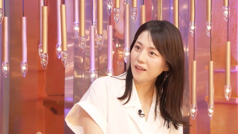 白いシャツを着た女性が、スタジオのような場所で、マイクを付けて穏やかな表情で話している様子