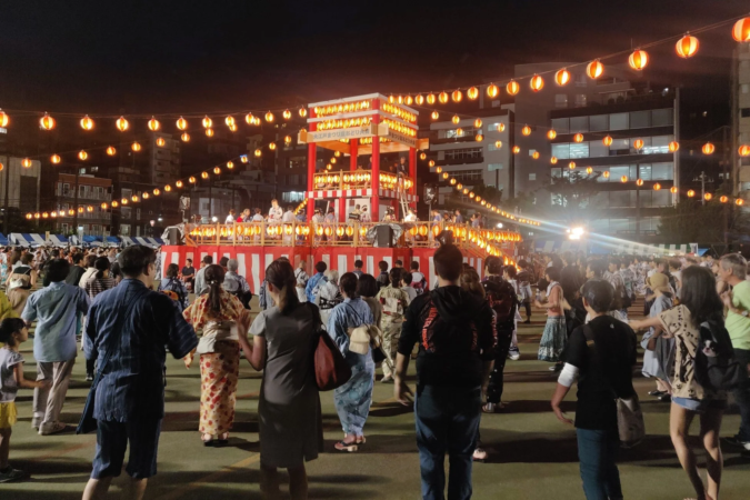 夜の盆踊り大会で、多くの人々が櫓を中心に踊っています。提灯が会場を明るく照らし、賑やかな夏の祭りの雰囲気を醸し出しています。