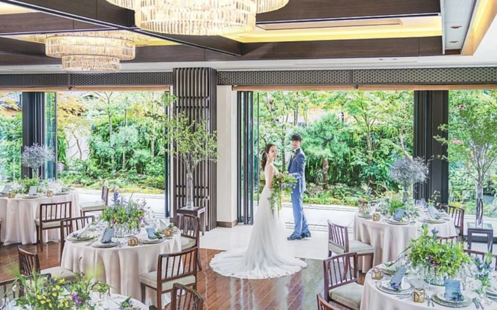 緑豊かな庭園を望む明るい披露宴会場で、ウェディングドレスとタキシード姿の新郎新婦が佇んでいます。