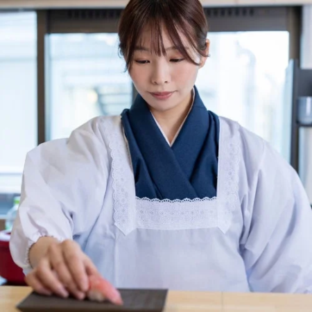 和服のような白い作業着を着た女性が、ネタを乗せた握り寿司を皿に置いている様子