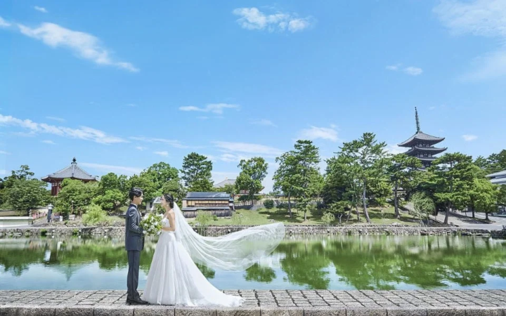 青空の下、池のほとりでウェディングドレスとタキシード姿のカップルが前撮り撮影をしている様子です。