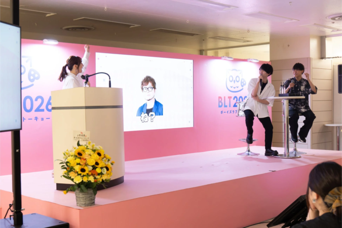 ピンクのステージで行われているイベントの様子。女性が演台で話し、スクリーンには男性の顔が表示されている。右側では2人の男性がマイクを持って座り、パネルディスカッションを行っているようだ