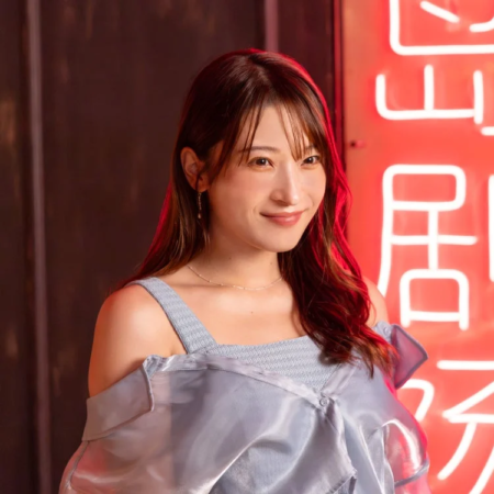 水色のオフショルダートップを着た笑顔の若い女性が、赤いネオンサインを背景にポーズをとっています。