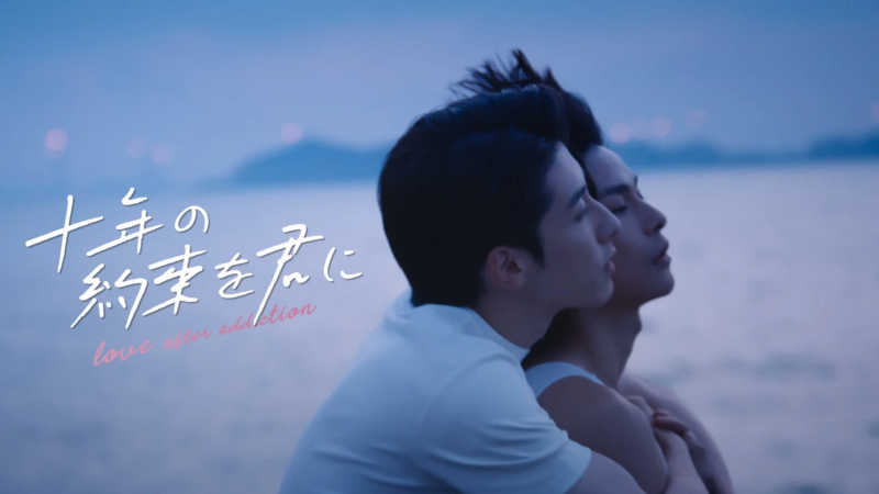 夕暮れ時の水辺で、二人の男性が抱き合い、お互いを優しく見つめ合っている