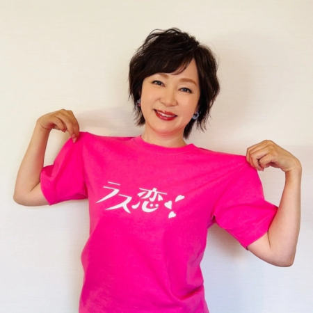 笑顔の女性が、白い壁を背景に「ラス恋！」と書かれた鮮やかなピンクのTシャツを着ています。