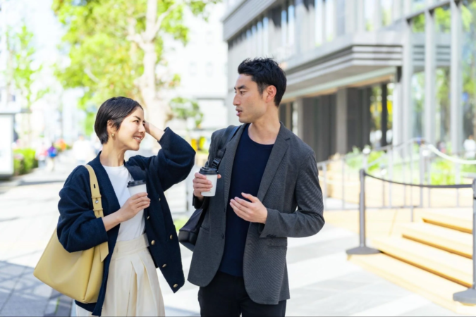 晴れた日の街中で、男女二人がコーヒーカップを片手に談笑しながら歩いています