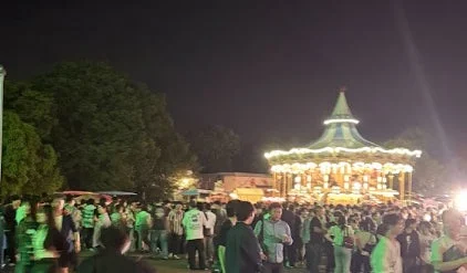 夜の遊園地で、多くの人々が賑わう様子を捉えた画像です。ライトアップされたメリーゴーランドが中央にあり、周囲は人でいっぱいです。