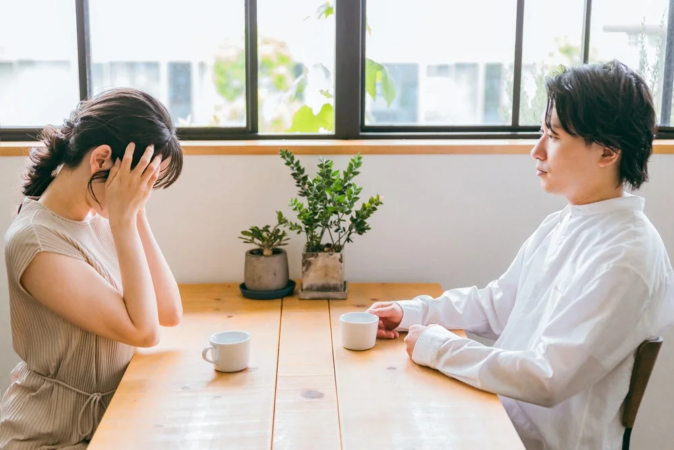 夫婦がテーブルを挟んで座っており、女性は顔を覆って悲しげな様子。男性は女性から視線を外し、二人の間に緊張感が漂っている。