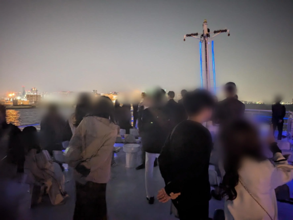 夜の船上デッキで人々が集まり、水辺に広がる街の夜景を楽しんでいる様子
