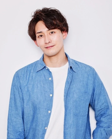 白い背景の前で、青いシャツと白いTシャツを着た若い日本人男性が優しく微笑んでいます。短めの茶色の髪で、カメラをまっすぐ見ています。
