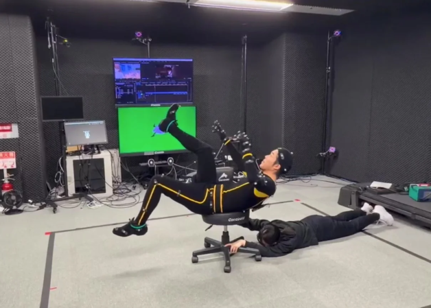 モーションキャプチャースタジオで、モーションキャプチャースーツを着た人物が椅子に座り、足を上げたユニークなポーズを取っている