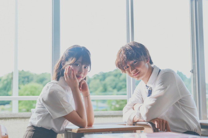 教室の窓際に座る若い男女の学生が、明るい自然光の中でカメラに笑顔を向けています